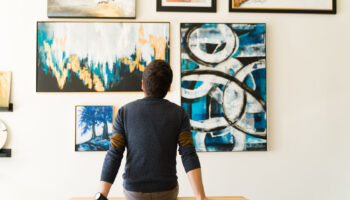 Hispanic male visitor looking reflective while sitting on a bench and admiring the various paintings on the wall of an art gallery